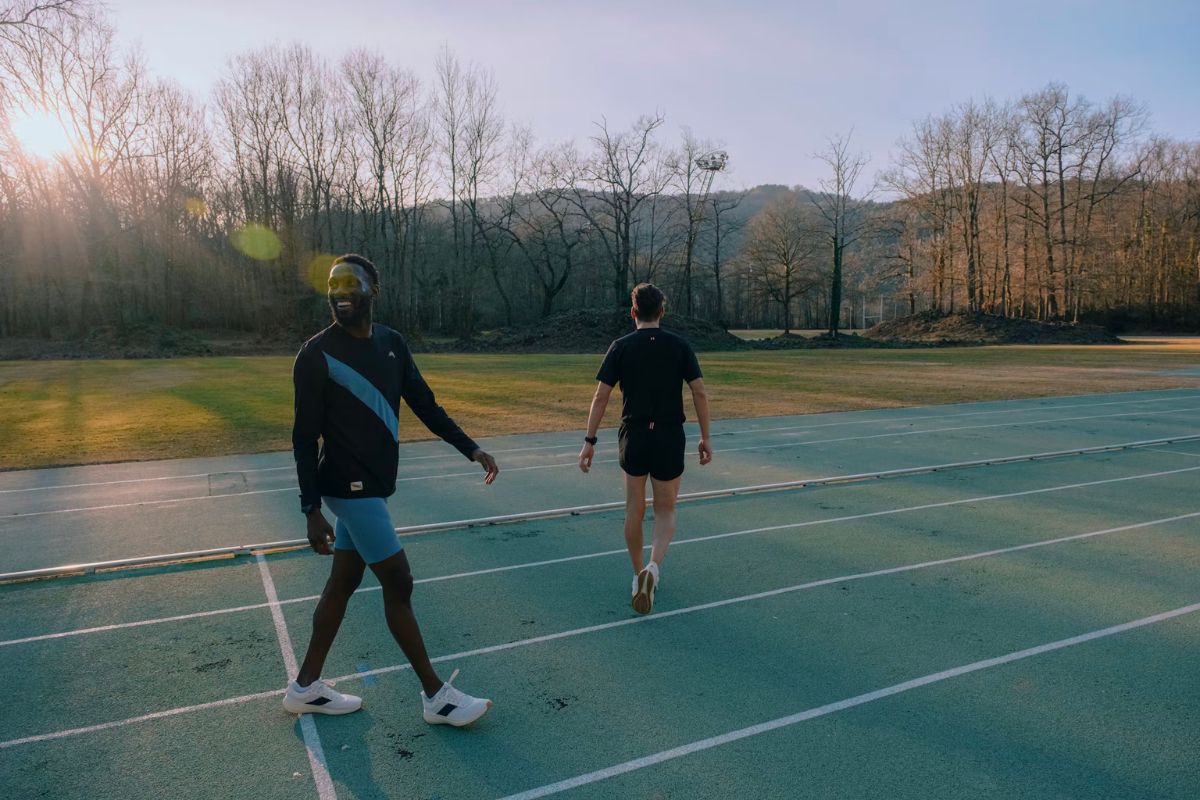 man smiling walking on a track wearing white running shoes with another man who has his back turnred
