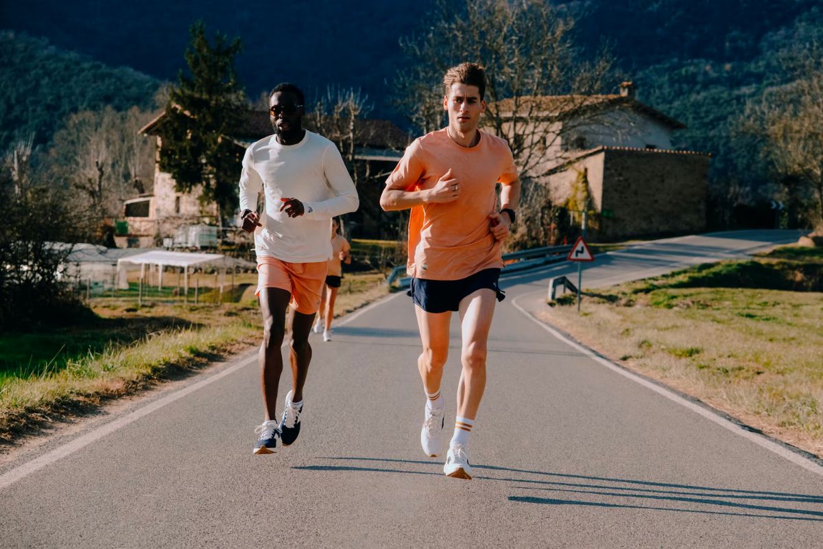 man in orange shirt running on a road with a man in a white long sleeve shirt running to the side behind him