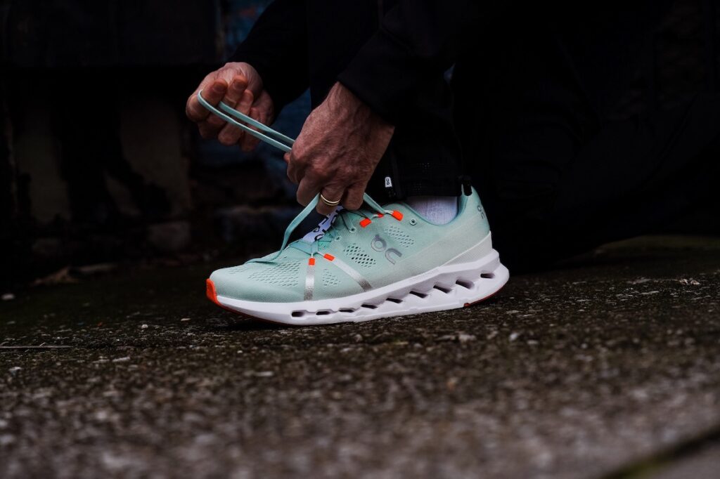 photo of hands tying shoelaces of a teal on cloudsurfer running shoe with orange accents