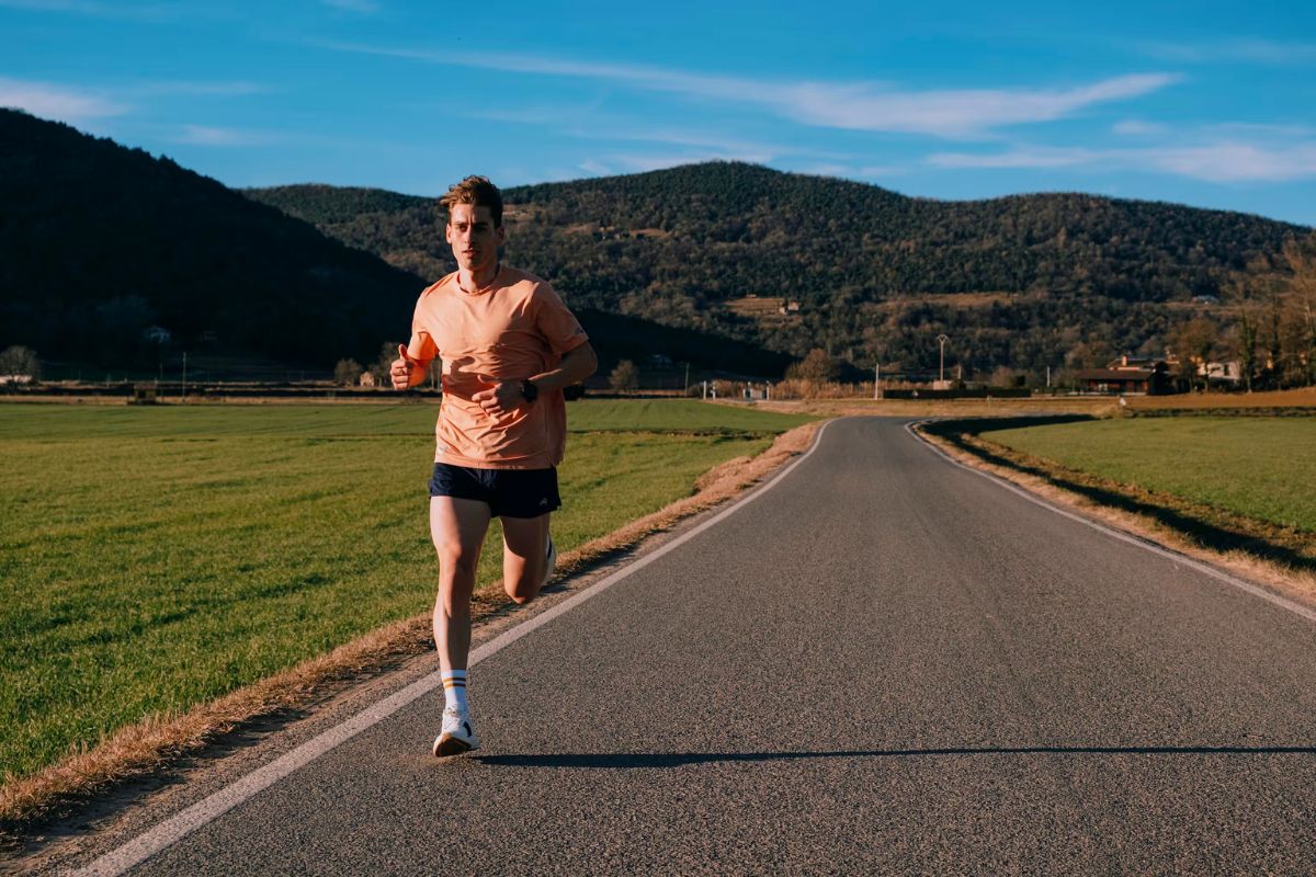 Man running in black shorts and peach colored shirt down an open road with mountains in the background