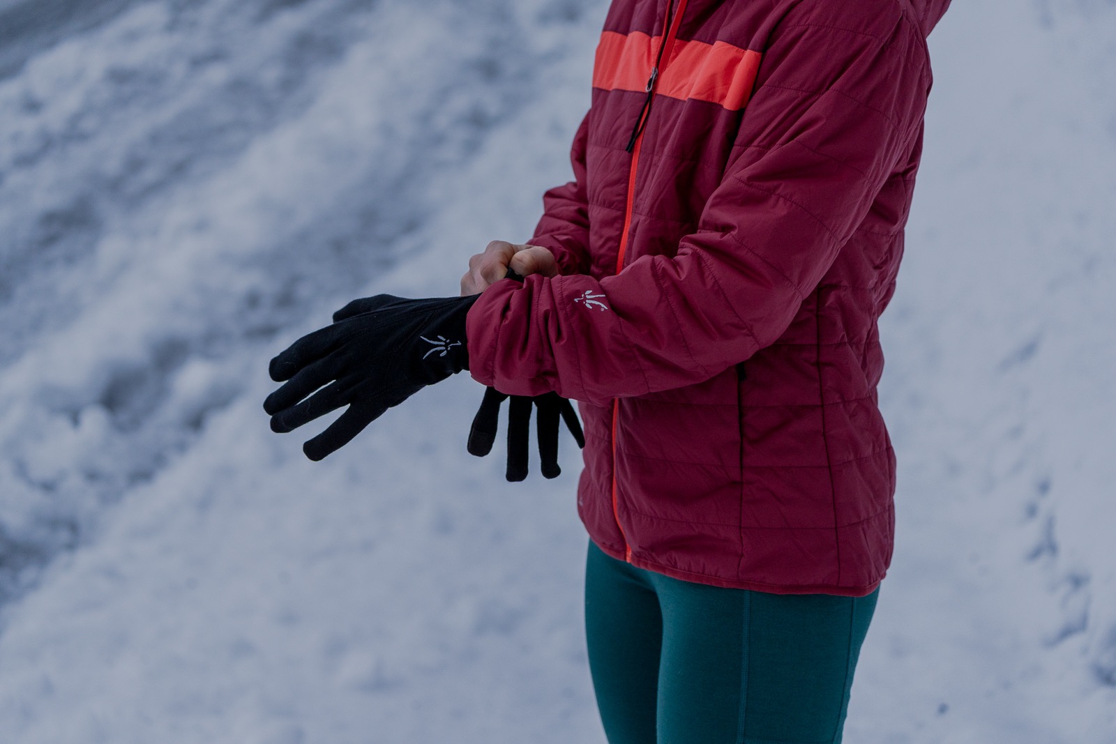 person wearing a red ibex wool aire hoodie on a snowy mountain putting on gloves