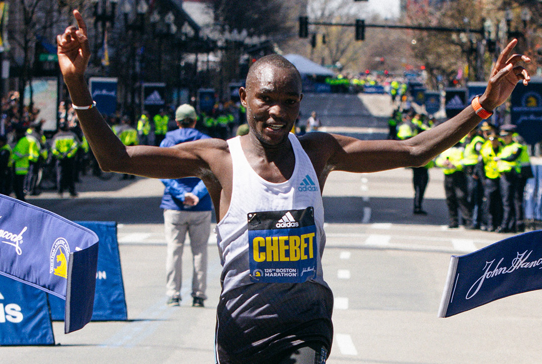evans chebet winning the boston marathon with arms raised in a white adidas singlet