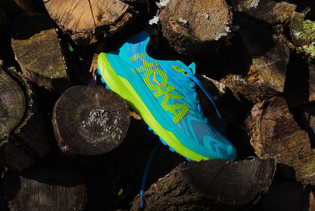 blue hoka trail shoe with yellow midsole on a pile of wood logs