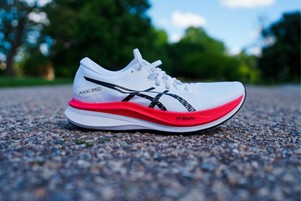 white asics shoe with red and white midsole on pavement with trees in background